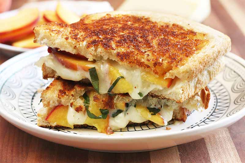 Two halves of a grilled cheese sandwich with peaches and basil stacked on top of each other on a white plate, with a plate of sliced fruit in the background, on a brown wooden cutting board.