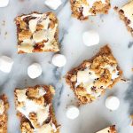 Top-down shot of marshmallow, chocolate, and graham cracker cookie bars, with scattered mini marshmallows on a gray and white marble surface.