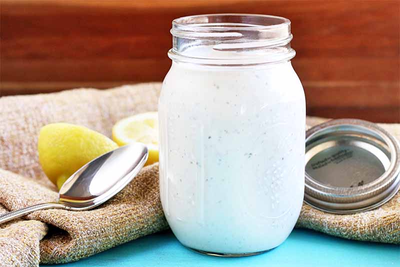 A mason jar of white barbecue sauce on a blue surface with a piece of brown burlap, a lemon cut in half, a spoon, and a lid, against a brown wood background.