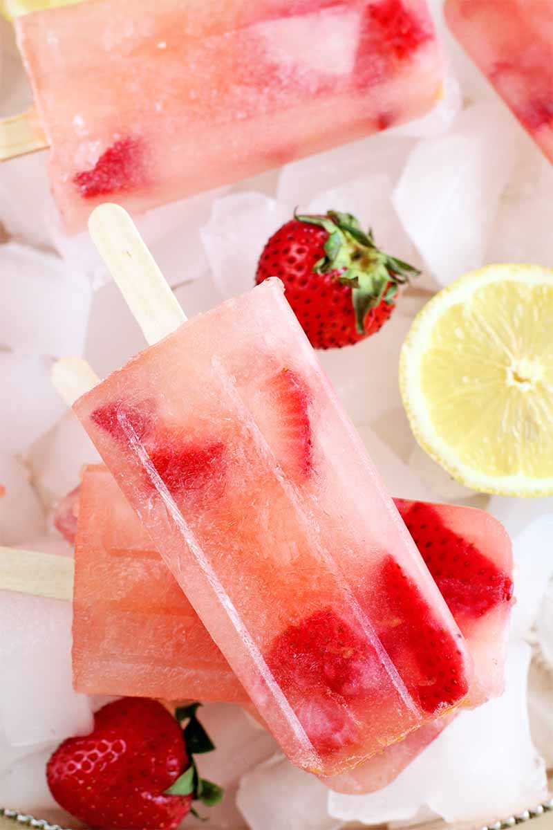 Pink strawberry lemonade popsicles are displayed on a bed of ice cubes, with whole fresh berries and yellow lemon slices.