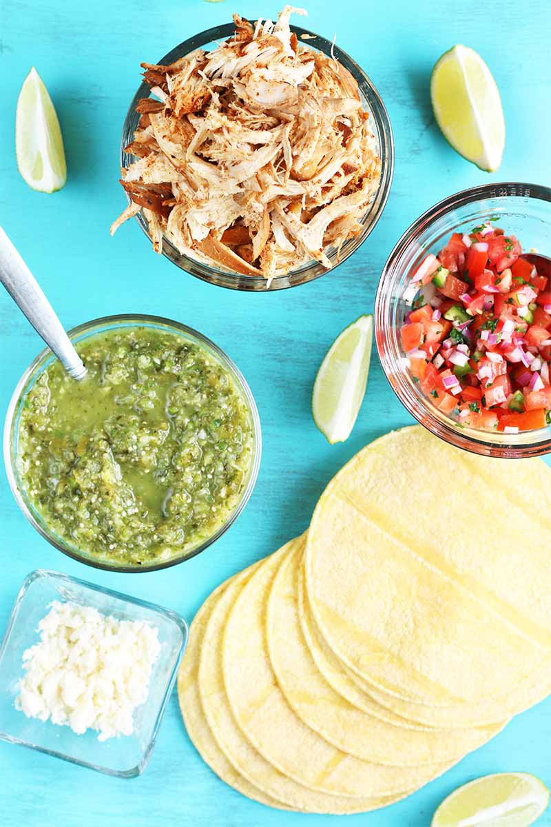Top-down shot of three clear glass bowls of grene salsa verde, spiced shredded chicken, and red pico de gallo, with lemon wedges, a small dish of chopped onion, and a fanned out stack of yellow corn tortillas, on a bright blue painted wood background.