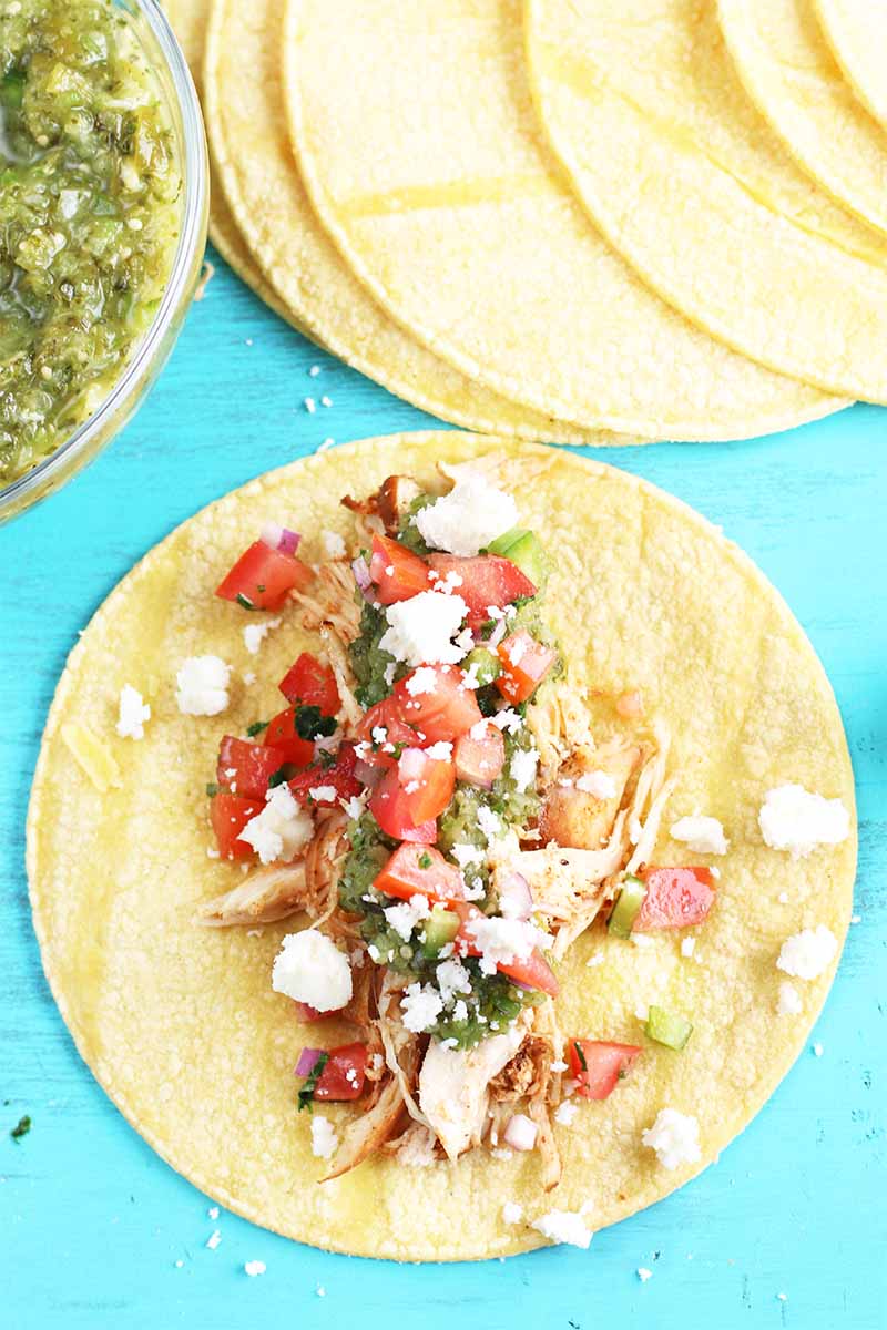 Top down shot of a corn tortilla topped with shredded chicken, crumbled cheese, and pico de gallo, with a fanned out stack of more yellow tortillas and a clear glass dish of green homemade roasted salsa verde, on a bright robin's egg blue background.