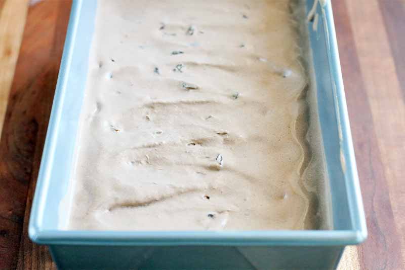 Oblique shot of a rectangular metal loaf pan filled with homemade ice cream, with a subtle zig-zag pattern on top, on a striped light and dark brown wood background.