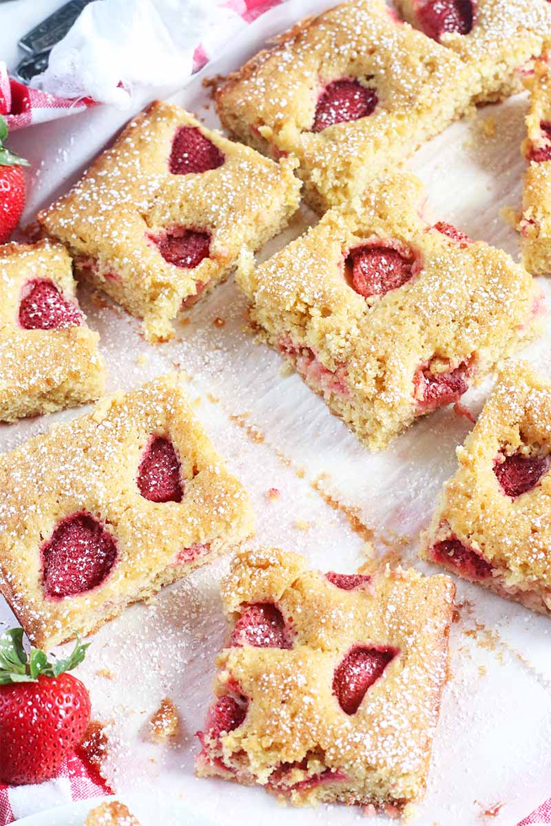 Square slices of strawberry cake arranged haphazardly on a white surface, with a few whole raw berries.