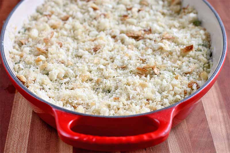 Fresh breadcrumbs top a vegetable mixture in a large oval white and red ceramic casserole dish with handles, on a brown wood surface.