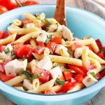 Tomato pasta salad in a blue bowl with a brown wooden spoon.