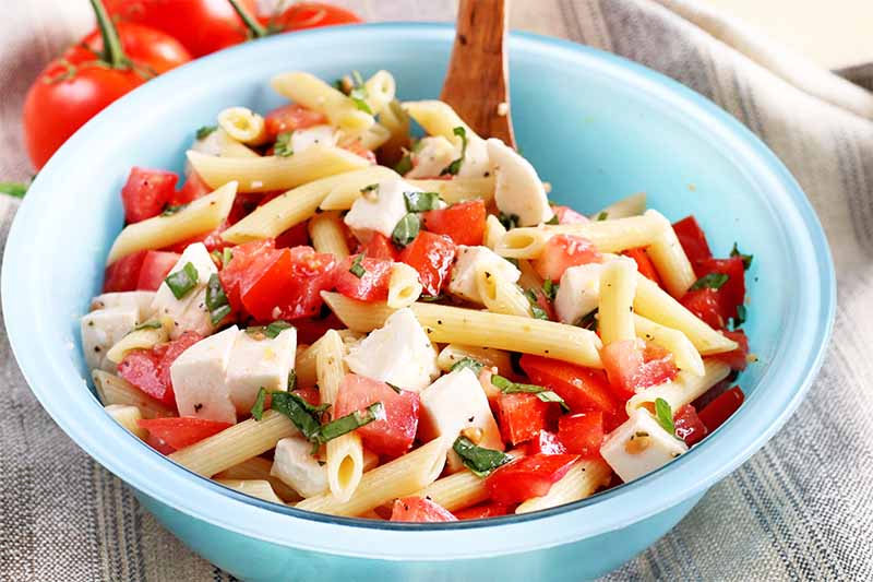 Tomato pasta salad in a blue bowl with a brown wooden spoon.