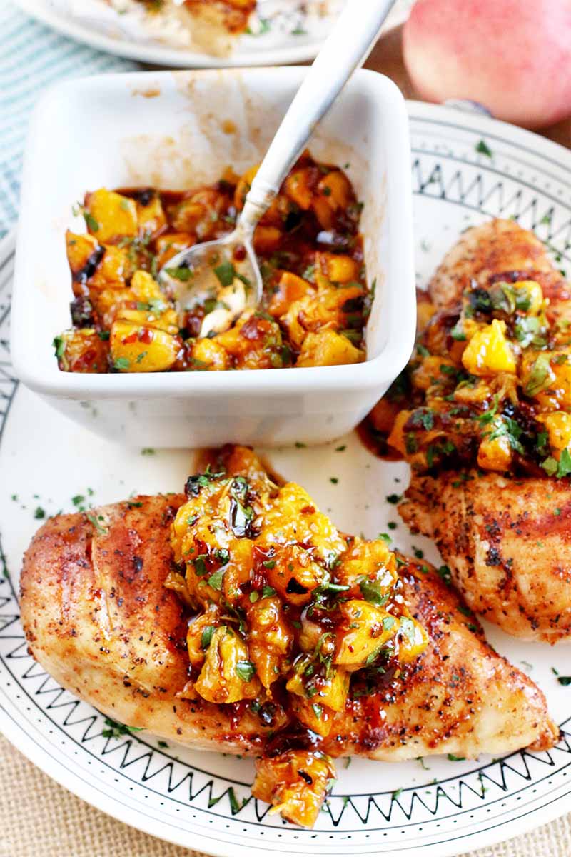 Two grilled chicken breasts beside a small dish of peach chipotle sauce with a spoon, with more on top of the protein, on a white patterned plate with a whole stone fruit and another dish in the background.