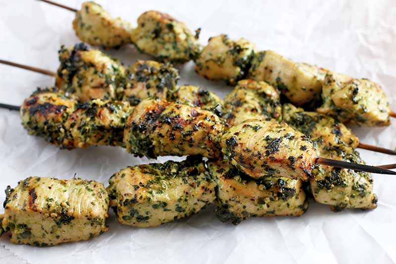 Four wooden skewers of grilled chicken, coated with an herb marinade, on a crumpled piece of white parchment paper.