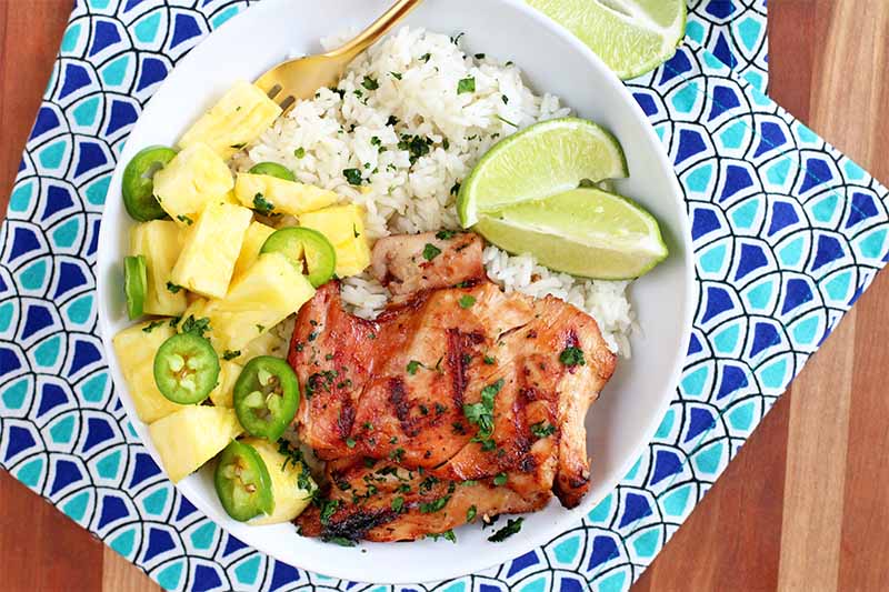 Top-down shot of a white bowl of grilled chicken, rice, pineapple, jalapeno, lime wedges, and fresh chopped herbs, on a dark and light blue patterned cloth, on top of a striped wood table.