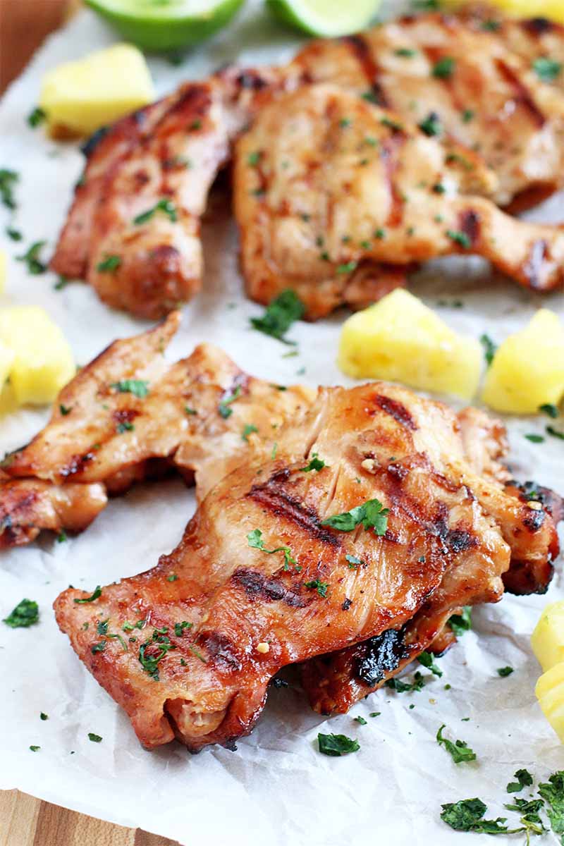 Two golden brown grilled chicken thighs on a piece of white parchment paper, with yellow pineapple chunks, chopped fresh herbs, and lemon wedges, on a brown wood surface.