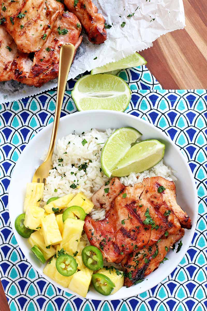 Top-down shot of a white bowl of white rice, lime wedges, pineapple chunks, jalapeno slices, chopped fresh herbs, and grilled chicken thighs, with a gold fork, on a blue patterned cloth with half of a green citrus fruit, and a piece of white parchment with more cooked poultry on it in the background.