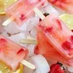 Pink strawberry lemonade popsicles with wooden sticks are arranged on a bed of ice cubes, with sliced lemon, and whole red berries with green tops.