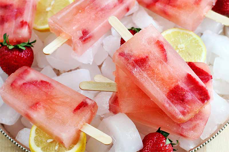 Pink strawberry lemonade popsicles with wooden sticks are arranged on a bed of ice cubes, with sliced lemon, and whole red berries with green tops.