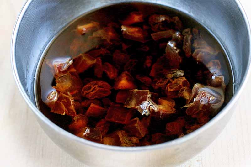 Raisins and chopped dates soak in spiced rum in a small stainless steel bowl, on a beige background.