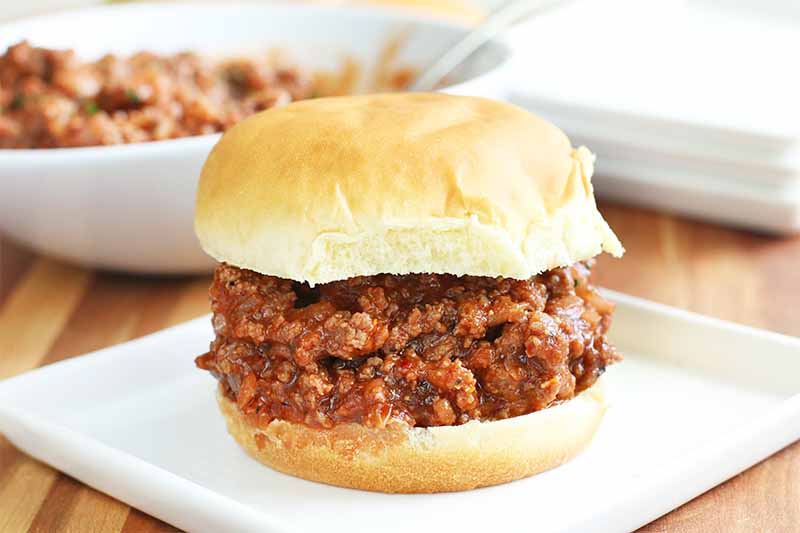 Sloppy joe ground beef sandwich with red sauce on a hamburger bun, on a white plate, with a bowl of more of the meat mixture and a stack of a few more plates in the background, on a brown wood surface.