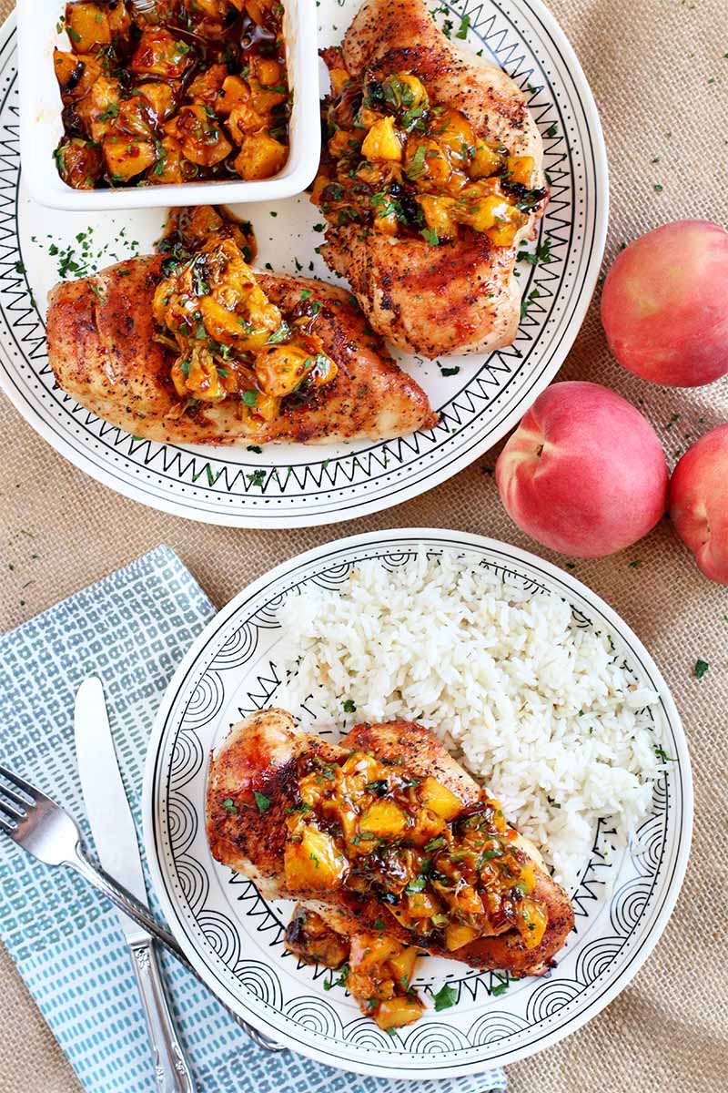 Top-down shot of two plates of grilled chicken, peach salsa, and white rice, with three whole stone fruit, a folded cloth napkin, a fork, and a knife, on a table topped with a piece of burlap.