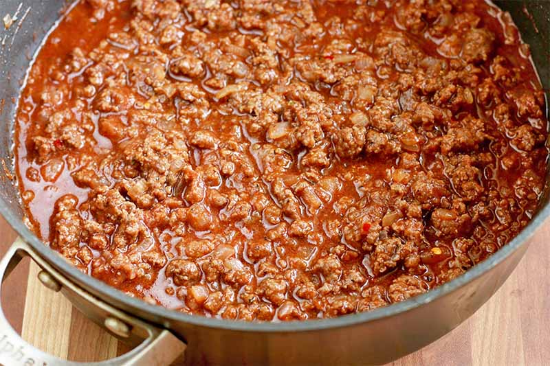 A large frying pan of ground beef in a red sauce, on a brown wood table.