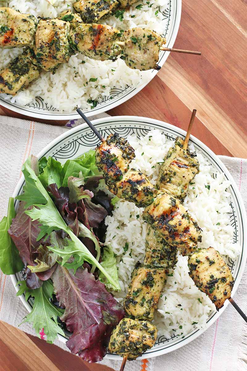 Two white dinner plates of honey lime chicken skewers, with white rice and a green salad, on a brown wood table covered partially with a white kitchen towel.