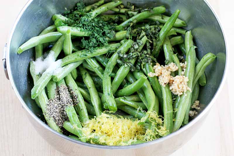 A stainless steel mixing bowl of fresh green beans topped with little piles of lemon zest, black pepper, salt, chopped green herbs, and minced garlic, on a beige background.