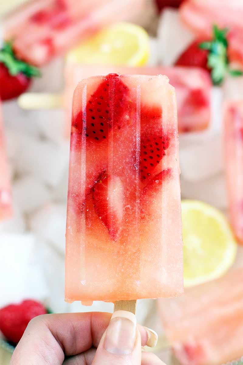 Vertical image of a French manicured woman's hand holding a strawberry lemonade popsicle up to the camera, with more of the frozen desserts along with whole berries and lemon slices on a bed of ice in the background.