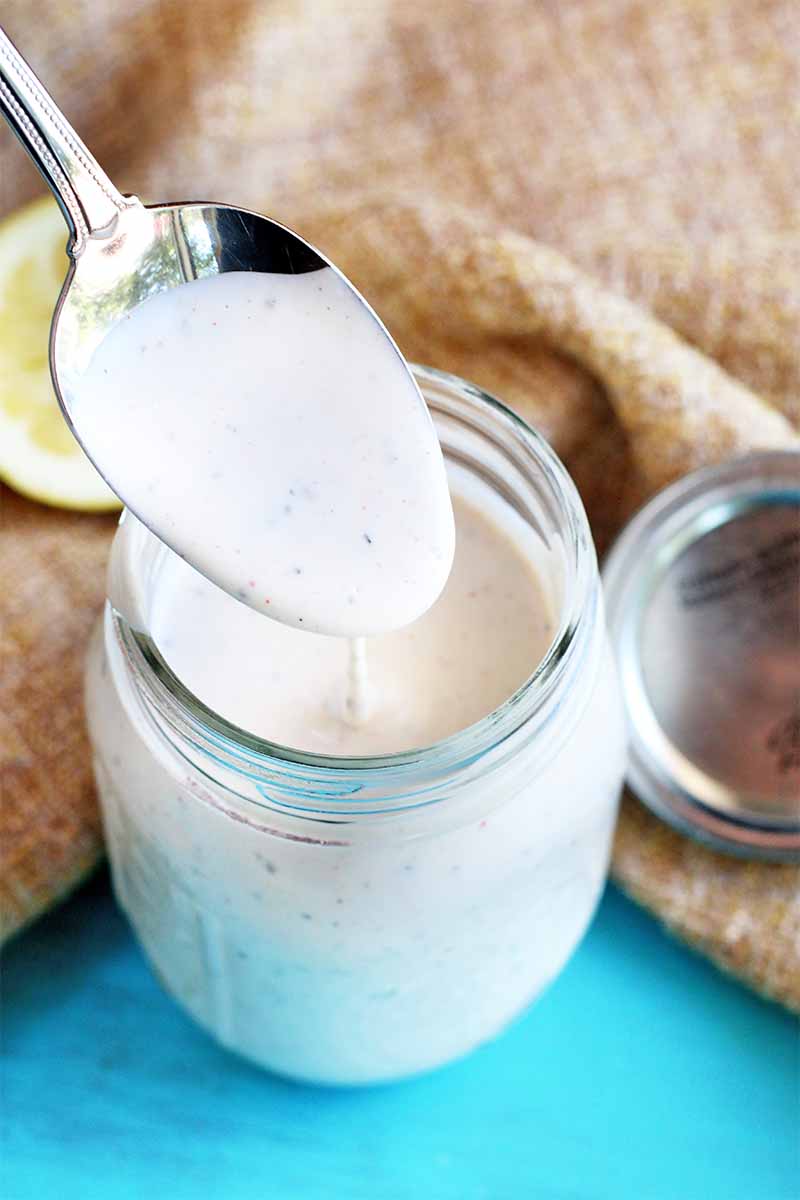 A spoon drizzles white barbecue sauce into a glass jar, with metal lid and half of a lemon on a piece of burlap, on a bright blue surface.