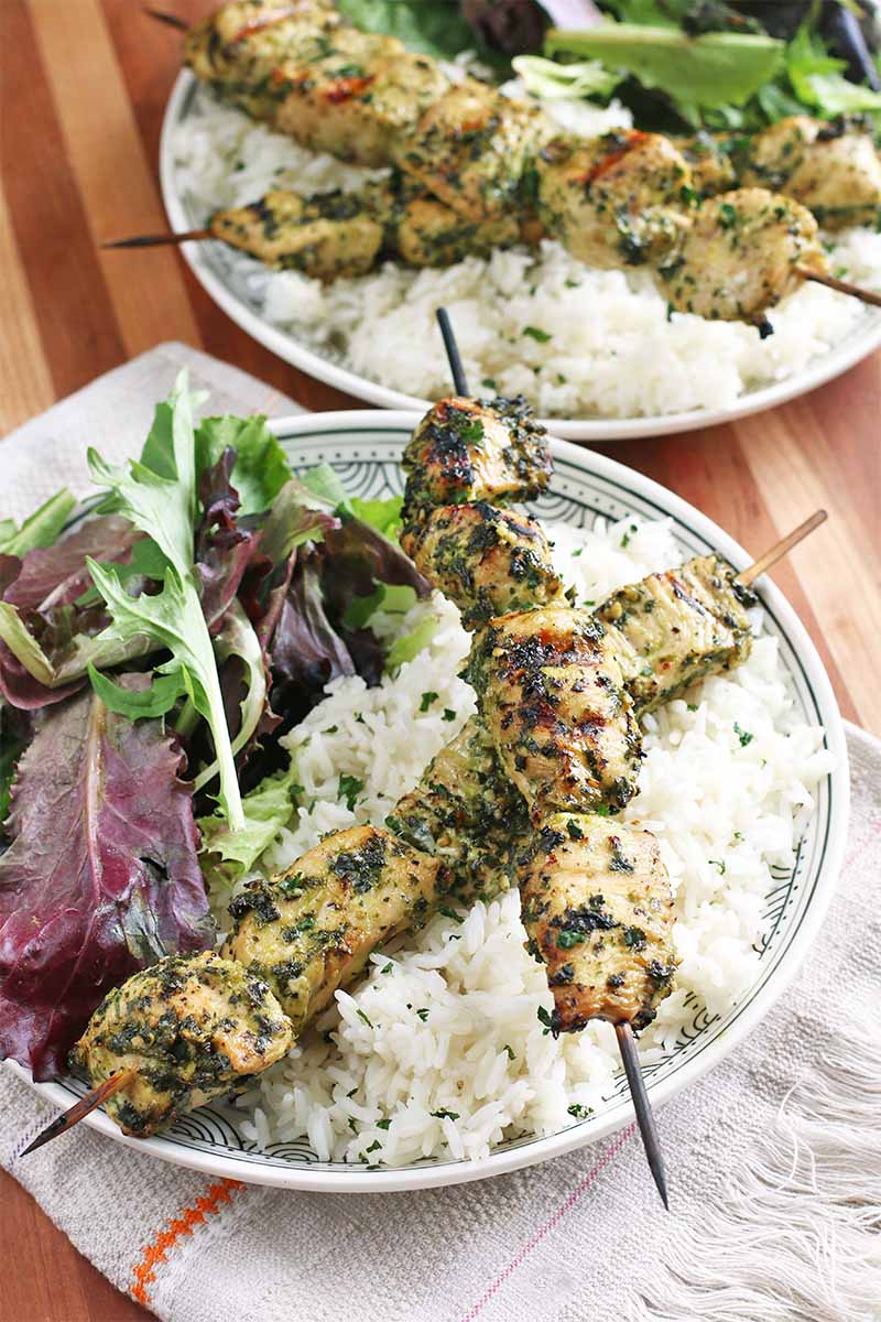 Two white dinner plates topped with two honey lime chicken skewers, white rice, and a green salad, with a white folded kitchen towel, on a brown wood table.