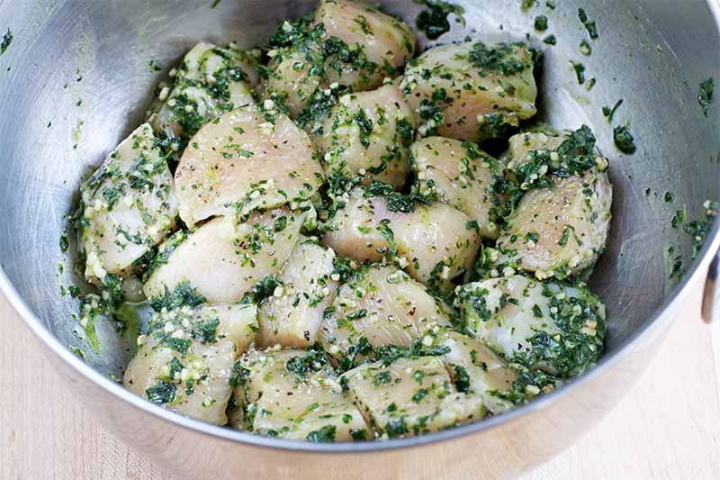 Raw chicken breast chunks tossed in a garlic and herb marinade, in a stainless steel bowl, on a beige background.