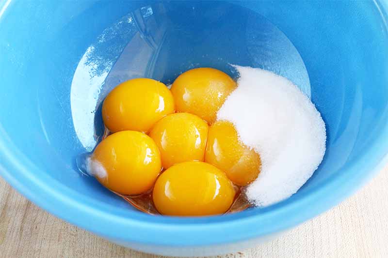 A blue glass bowl of six orange-yellow egg yolks and a pile of white granulated sugar, on a beige particle board background.