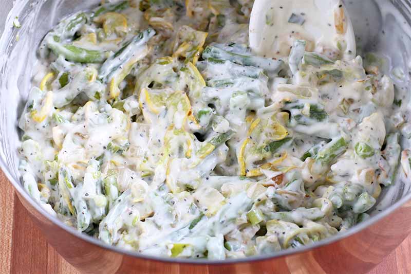 Roasted chopped yellow squash and green beans coated with a white cream sauce are being stirred together in a stainless steel mixing bowl with a wooden spoon, on a reddish brown wood surface.