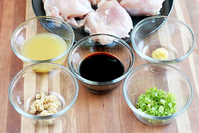 Five small glass dishes of pineapple juice, minced garlic, soy sauce, grated ginger, and chopped scallions, with a black plate of boneless skinless chicken thighs in the background, on a brown wood surface.