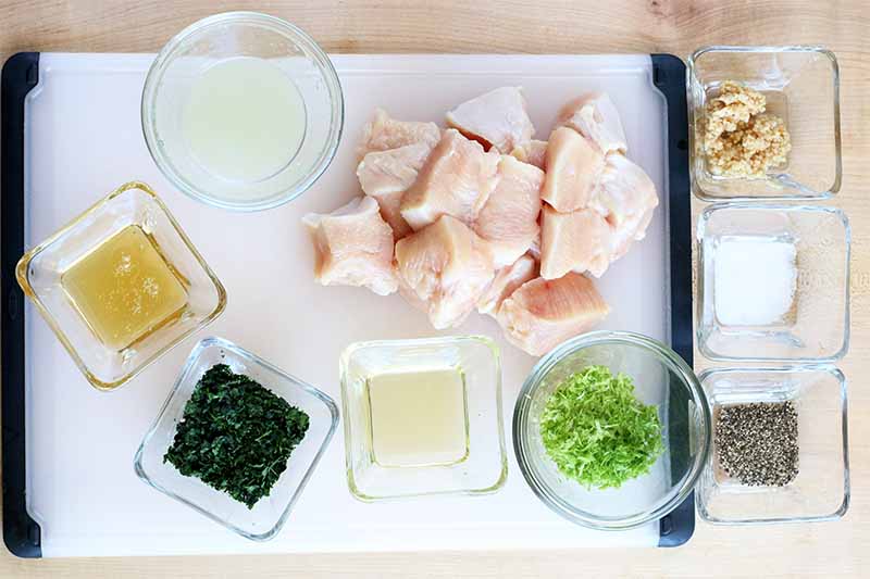 Top down shot of chunks of chicken breast, and small round and square dishes of minced garlic, salt, ground black pepper, lime zest and juice, chipped cilantro, canola oil, and honey, on a black and white plastic cutting board.