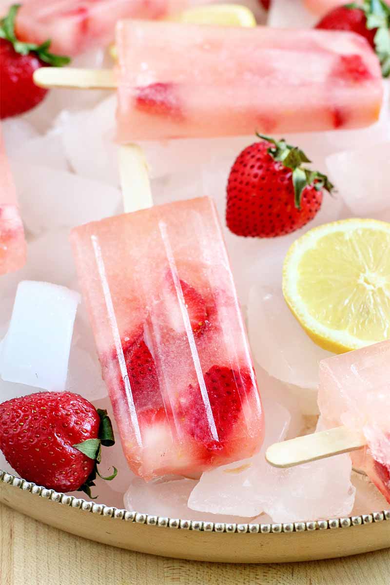 Pink strawberry lemonade popsicles arranged on a tray of ice, with whole fresh berries and lemon slices.