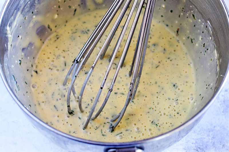 An egg and herb mixture in a stainless steel mixing bowl, with a wire whisk.