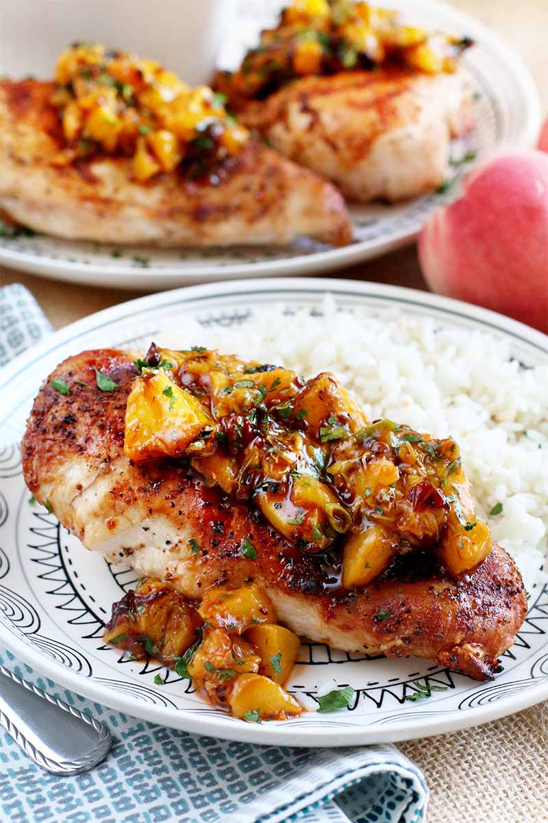 Two dinner plates of grilled chicken topped with a homemade peach salsa, with white rice, beside a whole stone fruit, a folded checked grayish blue and white cloth napkin, and a fork.