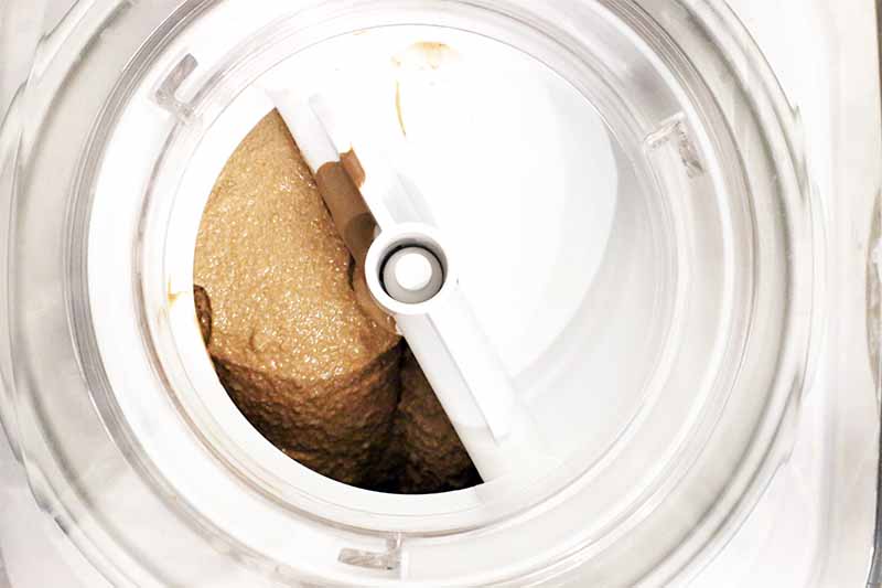 Top-down shot of a white and clear plastic Cuisinart ice cream maker, blending a brown chocolate mixture.