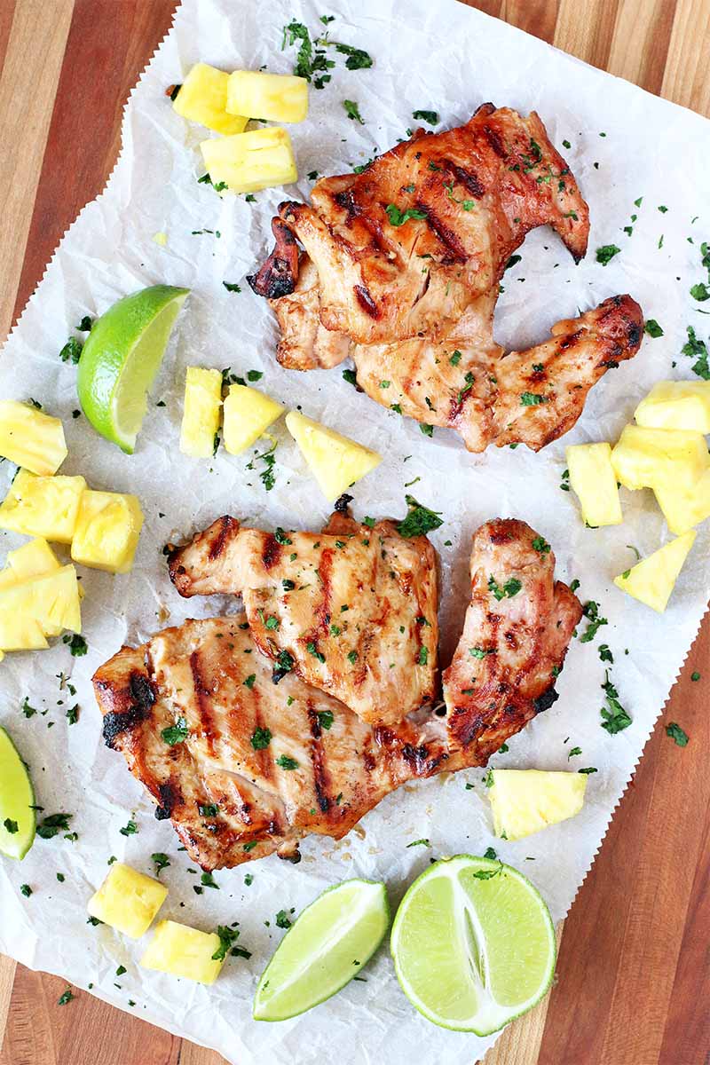 Top-down shot of two grilled chicken thighs on a piece of white parchment with pineapple chunks, lime wedges, and chopped green herbs, on a striped brown wooden surface.