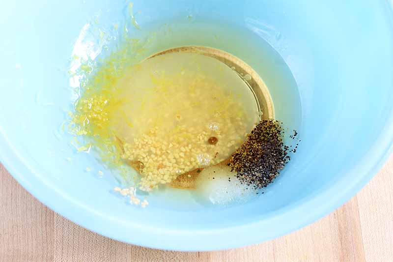 Black pepper, salt, lemon juice and zest, minced garlic, and olive oil in a blue glass bowl bowl on a beige surface.
