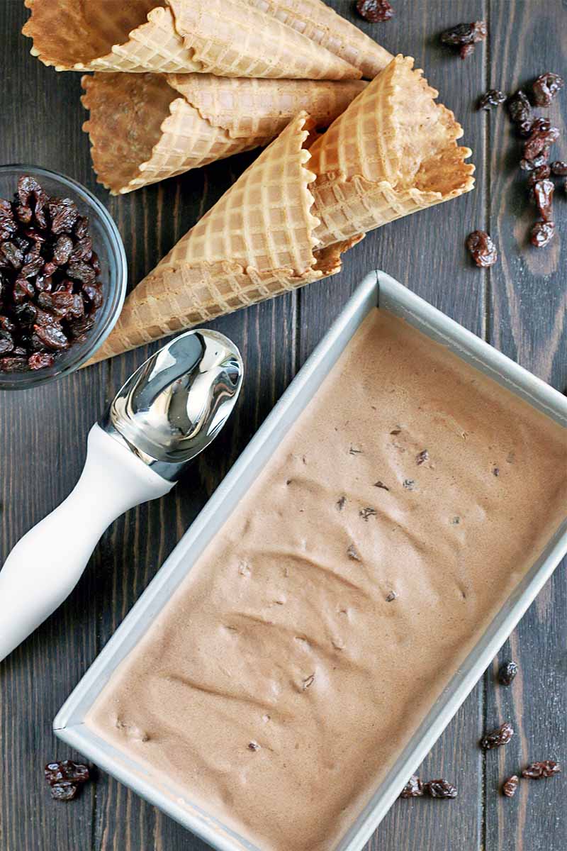 Top-down shot of a metal loaf pan filled with chocolate ice cream, next to a white and silver scoop, five waffle cones, and a small glass dish of brown raisins with more scattered around a dark brown wood surface.