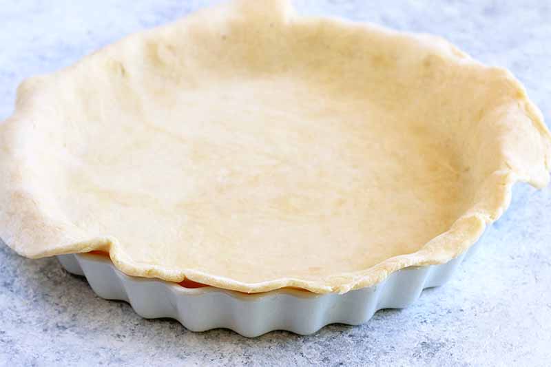 A white ceramic tart pan with scalloped edges is lined with dough.