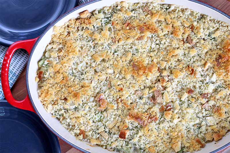 A red and white ceramic casserole dish is filled with a just-baked vegetable side topped with toasted breadcrumbs, with two dark blue plates in the background.