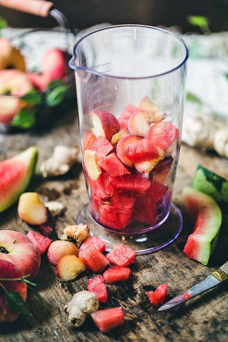A plastic blender canister filled halfway with chopped fruit, surrounded by more fruit, a paring knife, and watermelon rinds, on a brown wood surfac.