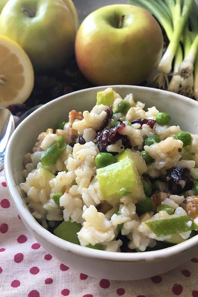 Vertical image of a bowl of rice salad with apples, green onions, and lemons in the background.