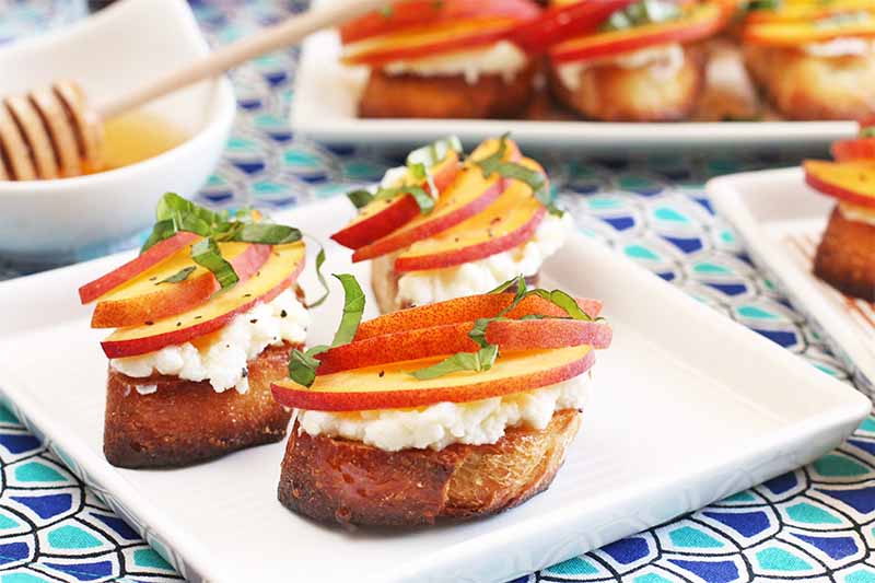 Three peach and basil crostini with ricotta on a square white plate, with twomore plates in the background, and a small round dish of honey with a wooden dipper, on a table topped with a dark and light blue cloth.