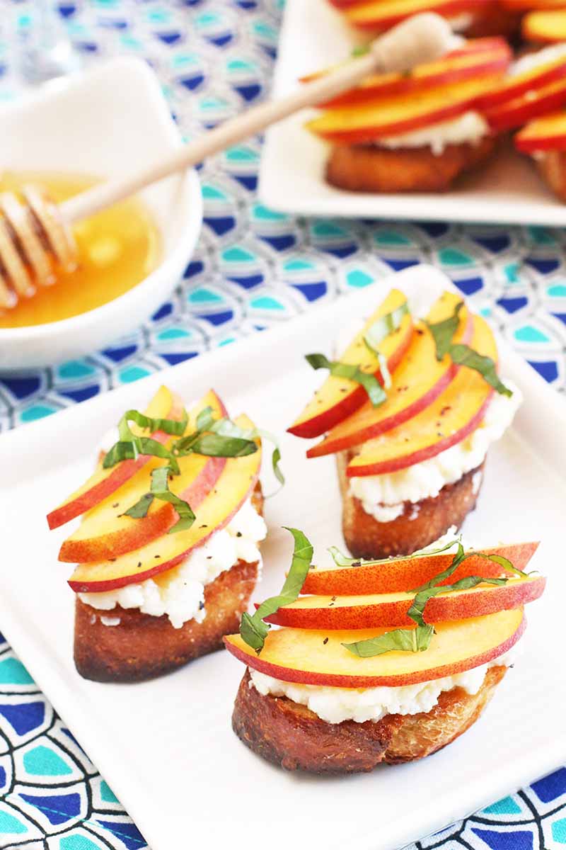 Three small baguette toast rounds topped with ricotta, honey, basil, and fresh peach slices, on a square plate with another identical plate and a small dish of honey with wooden dipper in the background, on a table topped with a light and dark blue and white patterned cloth.