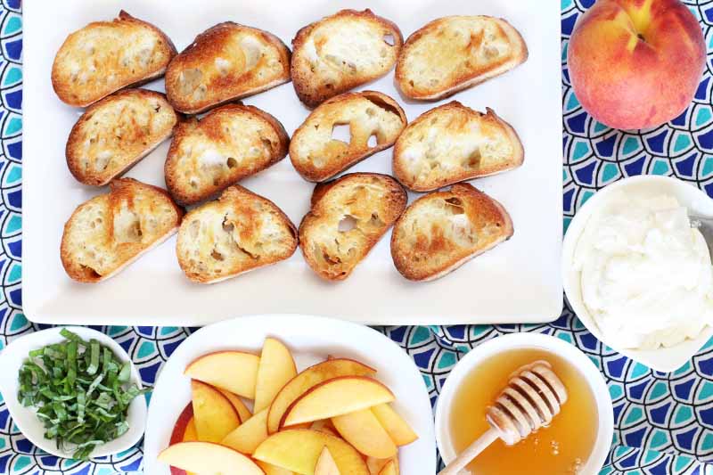 Top-down shot of a white rectangular serving dish topped with three rows of four toasted baguette slices, surrounded by a small dish of chopped basil, a bowl of sliced peaches and a whole fruit, a small bowl of honey with a wooden dipper, and a small dish of ricotta, on a light and dark blue patterned cloth.