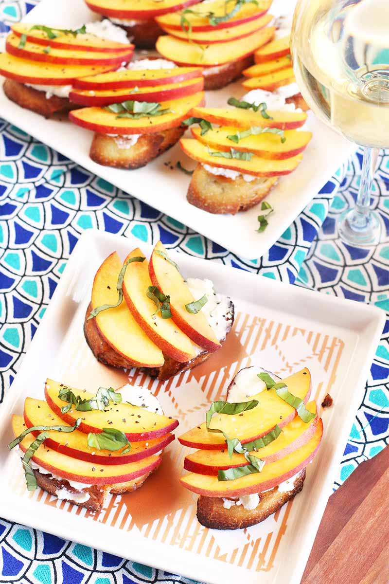 Two square plates of peach, basil, and ricotta crostini, on a blue patterned tablecloth, with a glass of white wine.