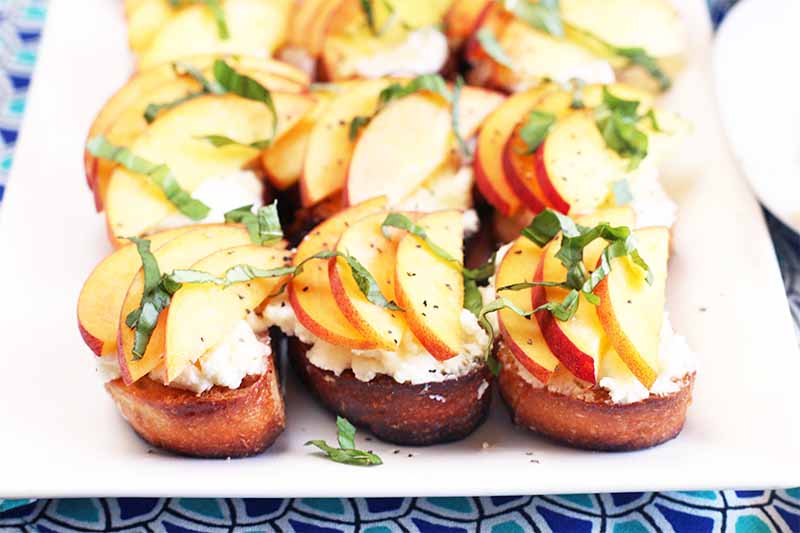 Three rows of three peach and basil ricotta toasts on a square white plate, on a table topped with a dark and light blue geometric patterned cloth.