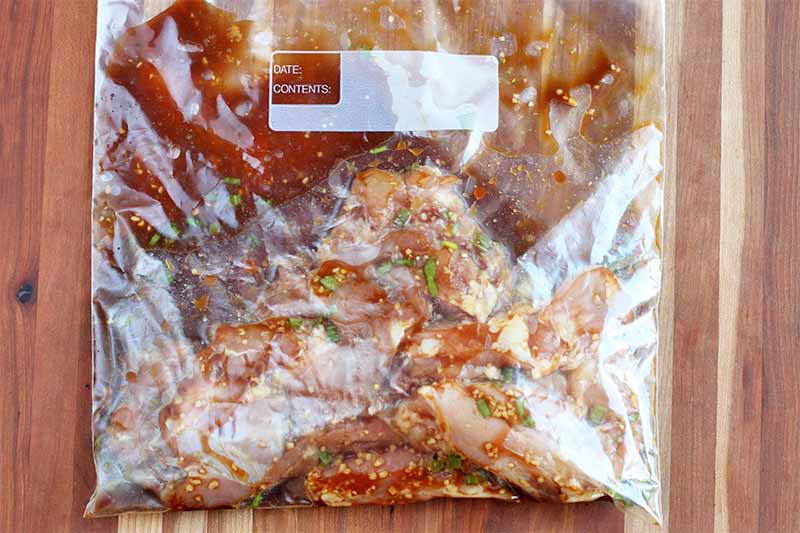 Raw chicken thighs in a brown marinade in a zip-top plastic bag, on a brown wood tabletop.