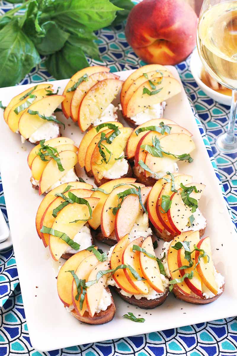 A white rectangular serving dish of twelve stone fruit and basil crostini with ricotta cheese, arranged in rows, on a blue patterned tablecloth with a sprig of mint, a glass of white wine, and a whole fruit.